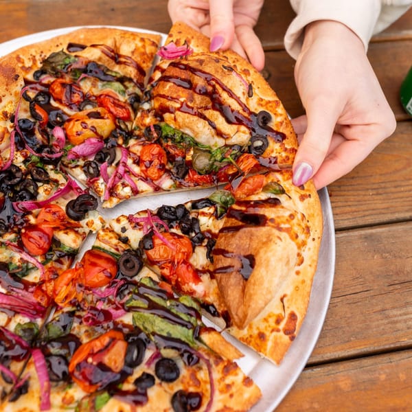 Hands taking a slice of MONDO PIZZA with tomatoes, olives, red onions, and balsamic glaze.