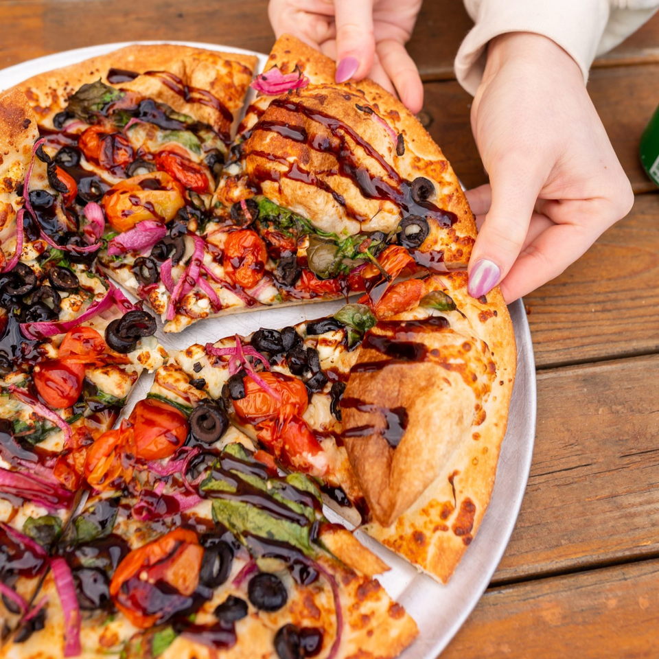 Hands taking a slice of MONDO PIZZA with tomatoes, olives, red onions, and balsamic glaze.