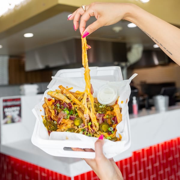 Hands pulling a string of melted cheese from loaded fries with jalapeños, bacon, and green onions.