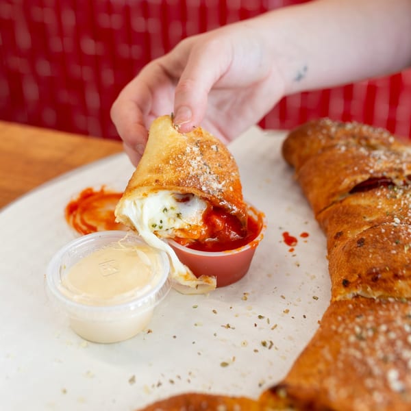 Hand dipping a cheesy calzone into a small red cup of marinara sauce at MONDO PIZZA.