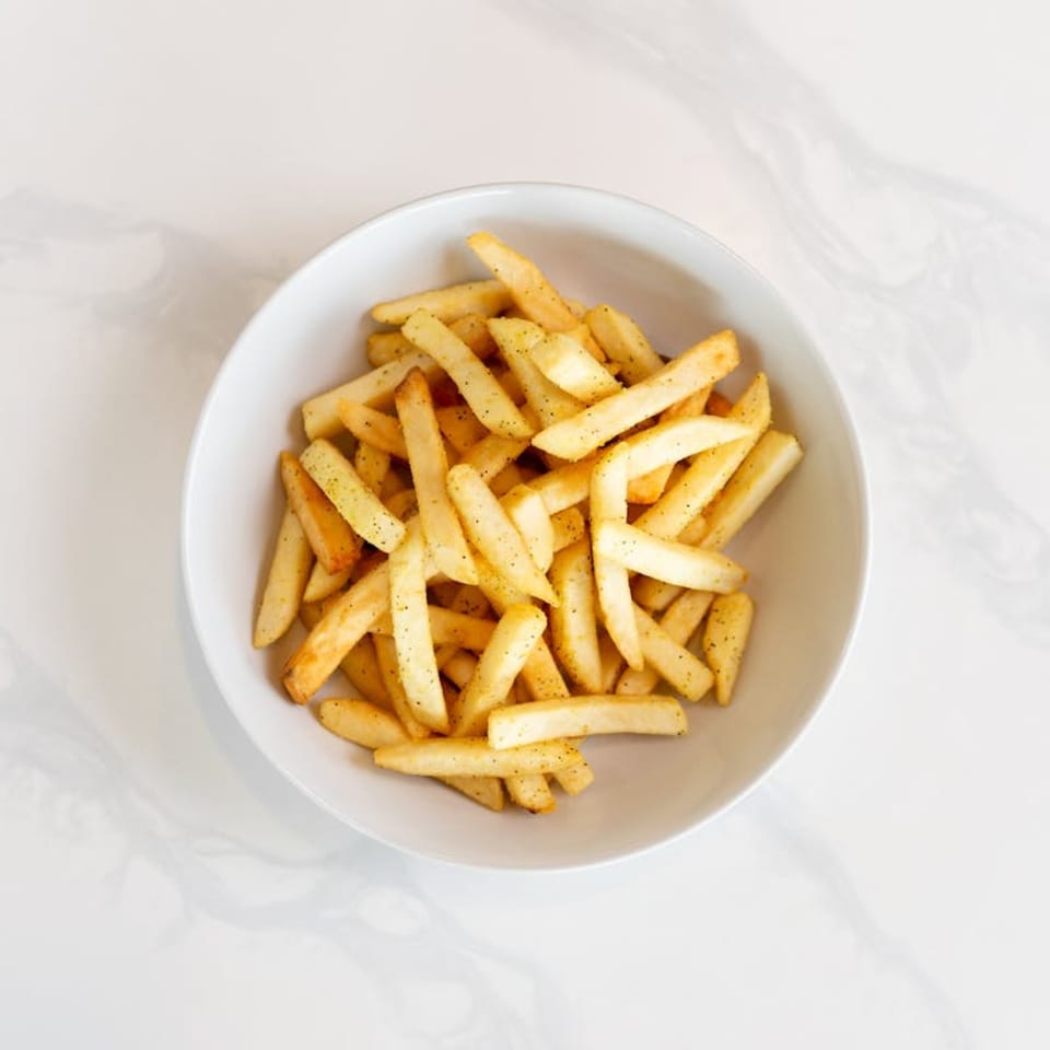 A white bowl filled with golden, seasoned french fries on a white marble surface.